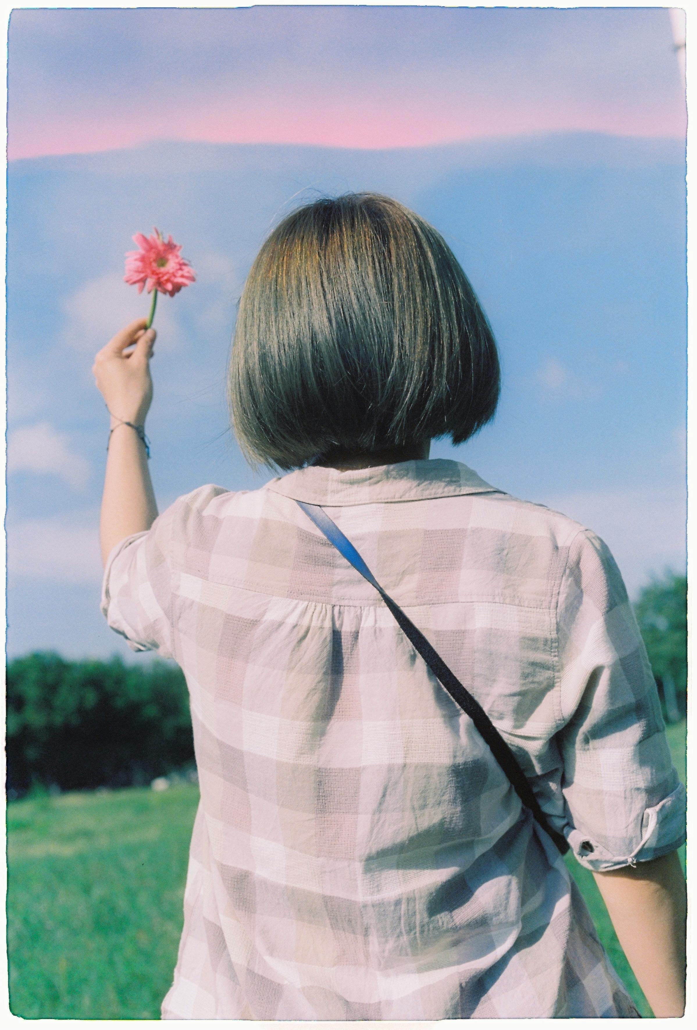 A young woman holding a flower in a scenic field under a blue sky with pink horizon.