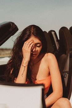 A thoughtful woman sits in a convertible car under the bright daylight, reflecting deeply.