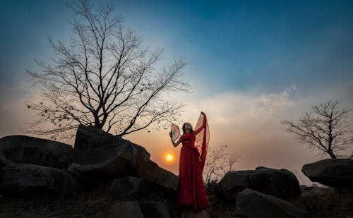 Woman in Red Dress with Hand Fan Posing on Rocks at Sunset