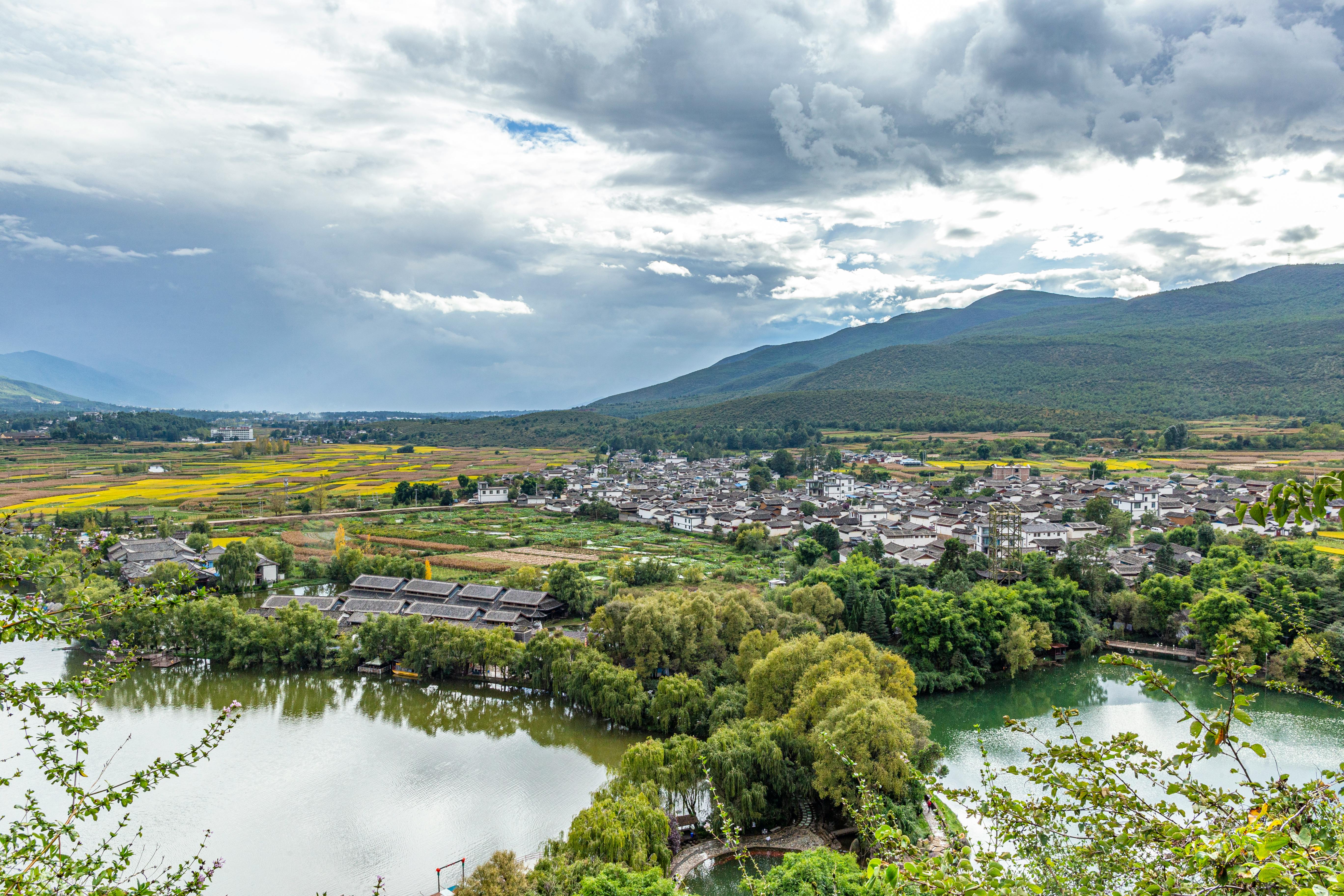Picturesque view of a mountain town with lush fields and a serene lake under dramatic skies.