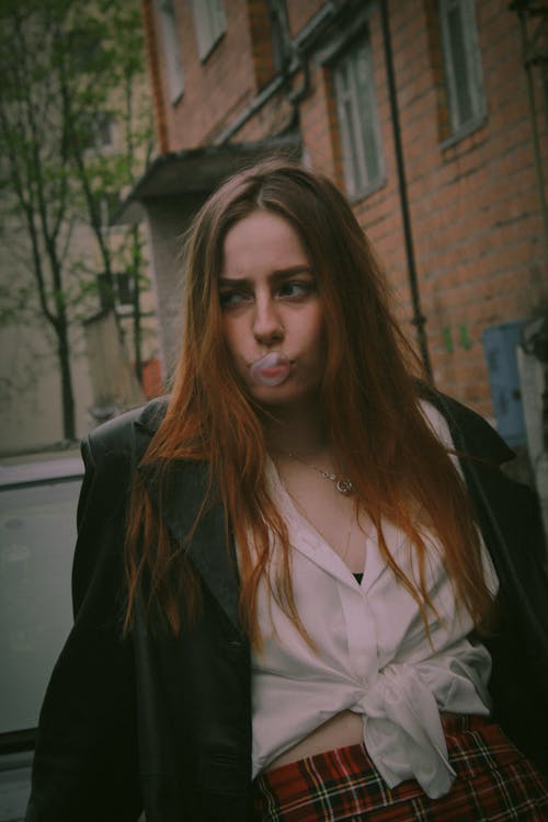 Young Woman in a White Shirt, Plaid Skirt and Black Coat Standing Outside and Making a Bubble Gum Balloon 