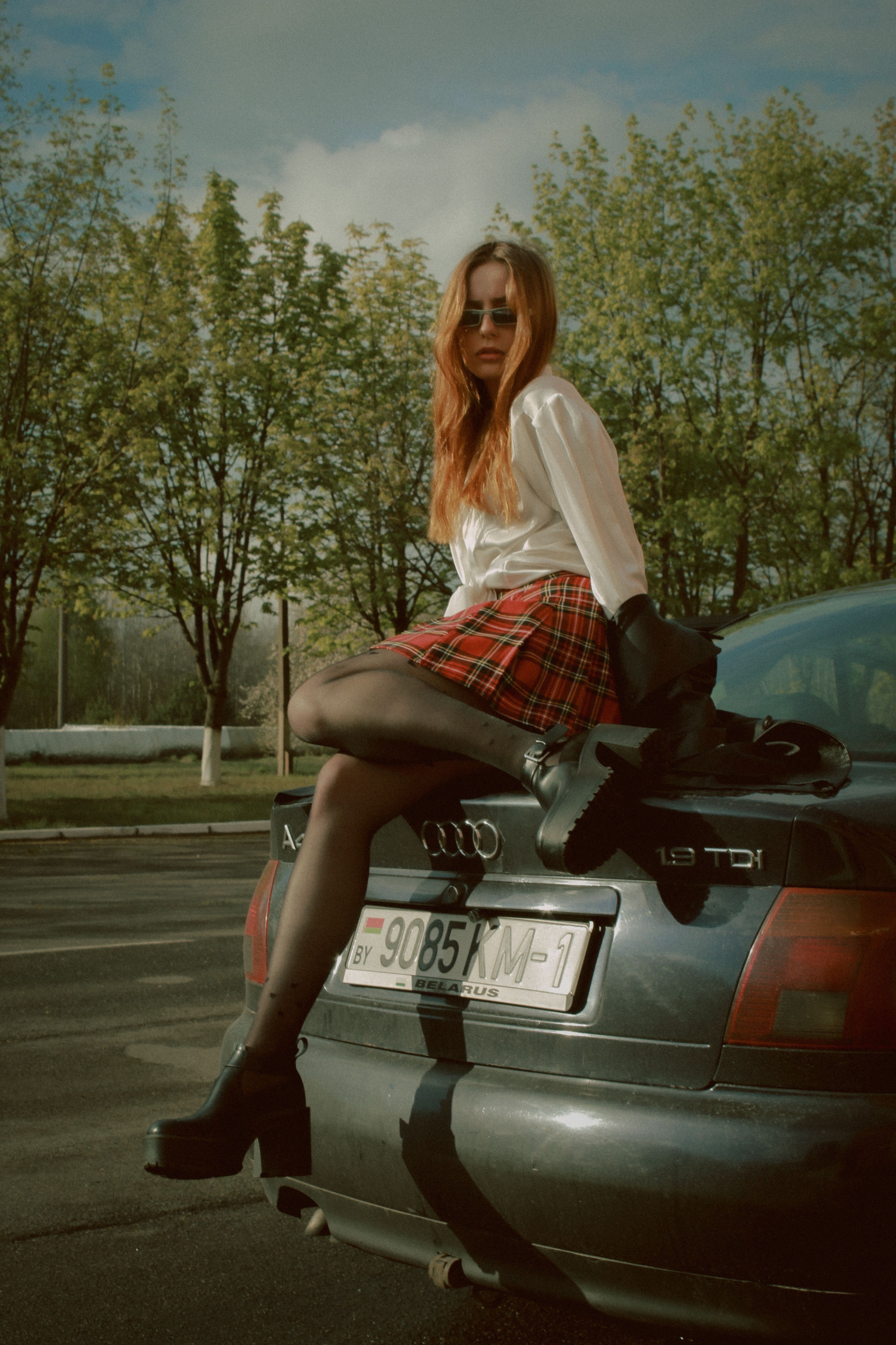Stylish woman in plaid skirt sitting on an Audi car outdoors, wearing sunglasses and boots.