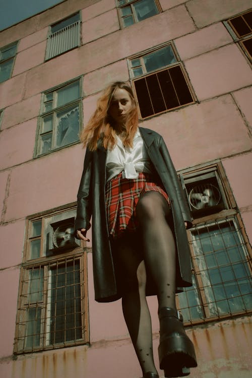 Young Woman in a White Shirt, Plaid Skirt and Black Coat Standing in front of an Abandoned Building 