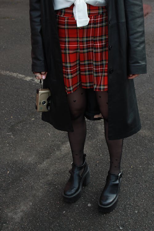 Woman in a Red Plaid Skirt and a Black Coat Standing on a Street and Holding a Camera