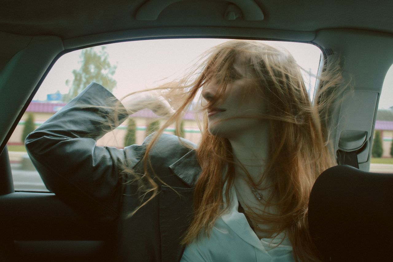 a young woman with red hair sits in a car smiling as her hair blows in