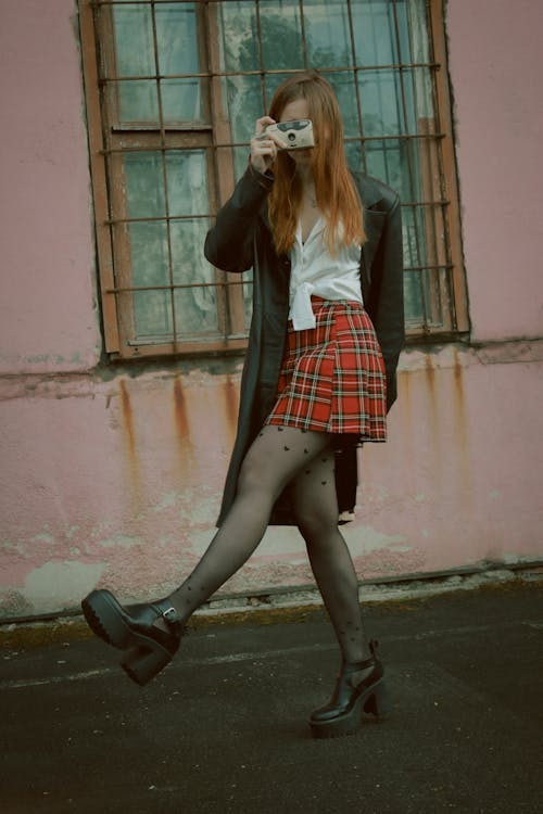 Woman in a Red Plaid Skirt, White Shirt and a Black Coat Standing in front of a Pink Building