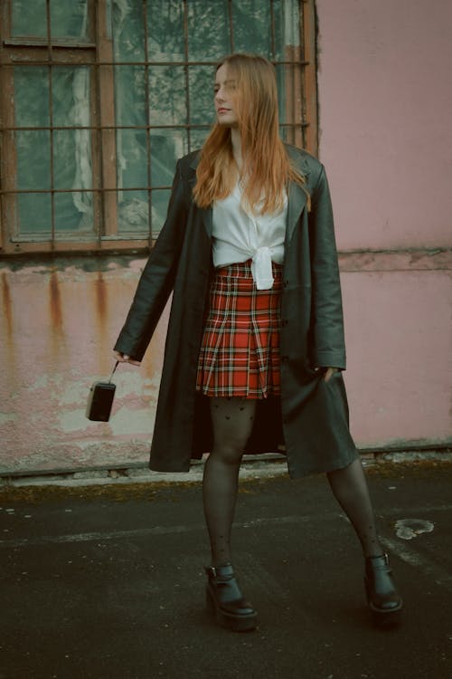 Woman in a Red Plaid Skirt, White Shirt and a Black Coat Standing in front of a Pink Building