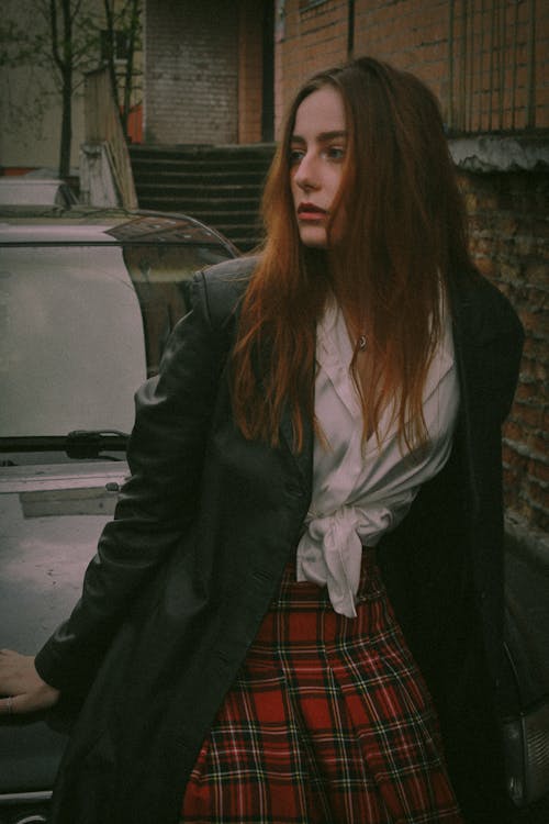 Young Woman in a White Shirt, Plaid Skirt and Black Coat Leaning against a Car 