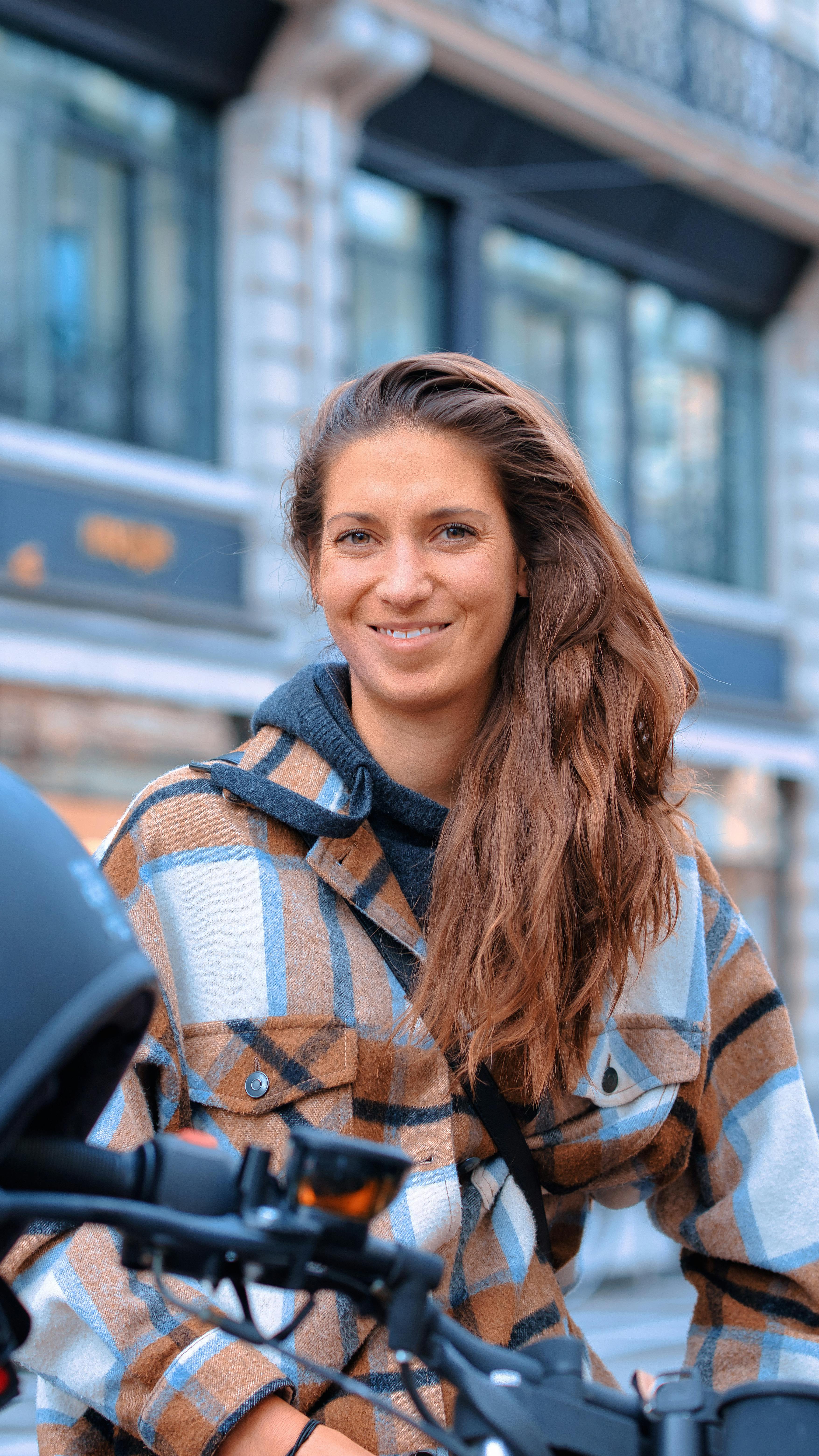 Smiling woman with brown hair poses outdoors in a plaid coat, embracing urban style.