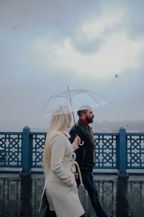 Free A couple walks under a transparent umbrella on a rainy, overcast day by the water. Stock Photo