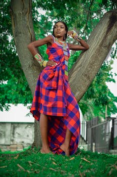 Elegant woman striking a pose in a vibrant checkered dress against a tree in Kinshasa park.