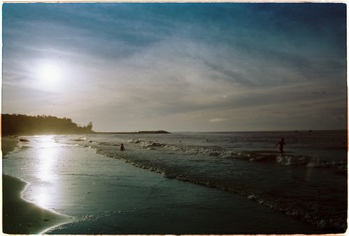 Free Serene beach scene at sunset with people enjoying the waves and clear sky. Stock Photo