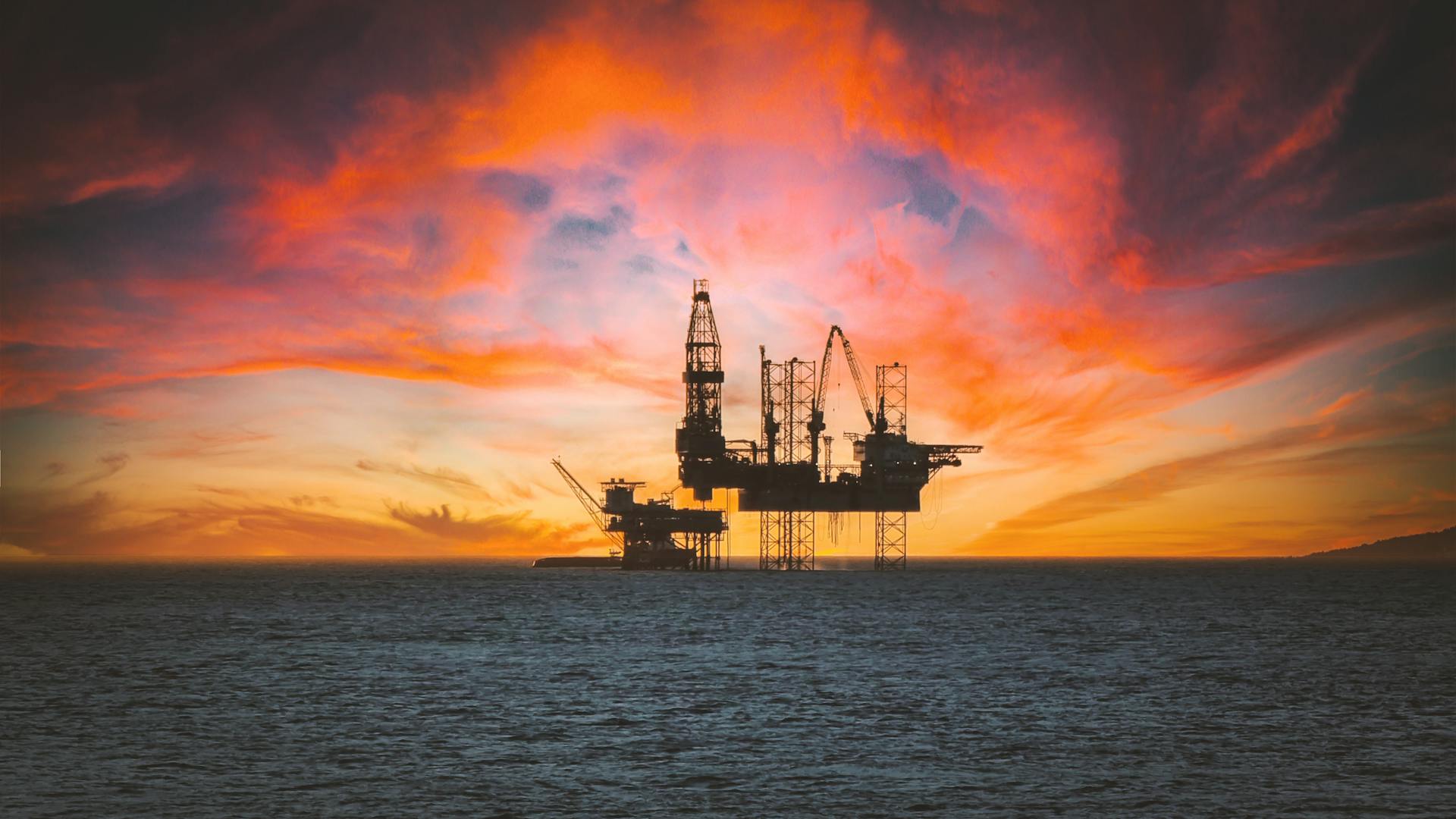 Offshore oil rig silhouetted against a vibrant sunset with dramatic clouds.