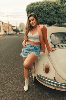 A fashionable woman in denim shorts poses confidently by a vintage car on a vibrant city street.