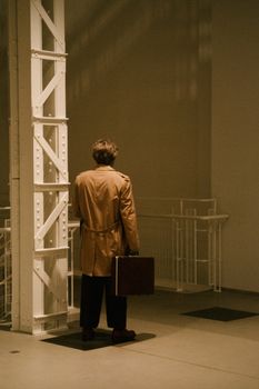 A person in a trench coat with a suitcase stands alone in a dimly lit corridor.