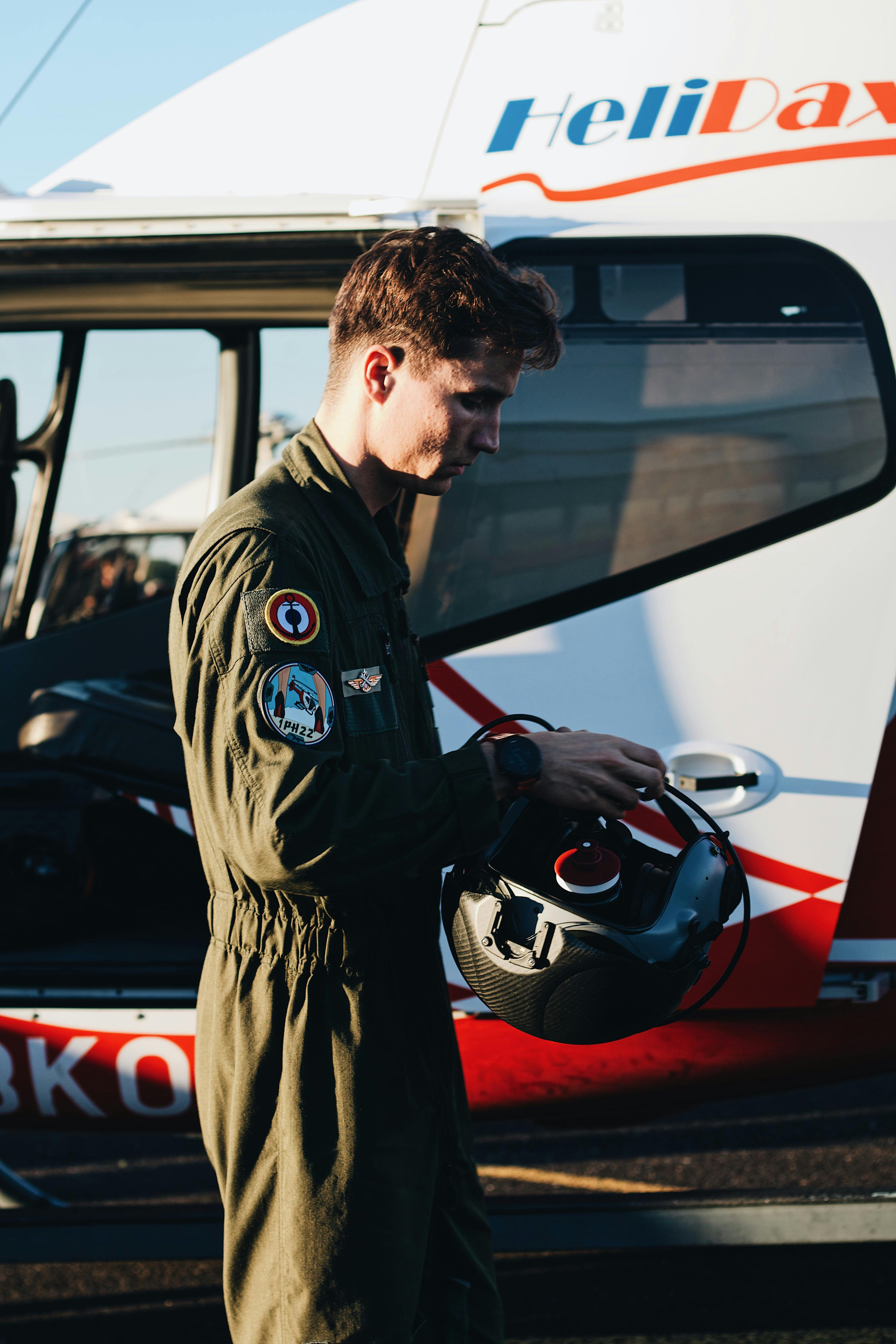 A pilot wearing a flight suit holds a helmet beside a helicopter on a sunny day.