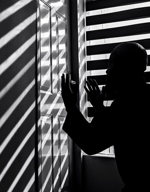 Window Blinds Casting Shadows on a Closed Door with a Silhouette of a Praying Man in the Foreground