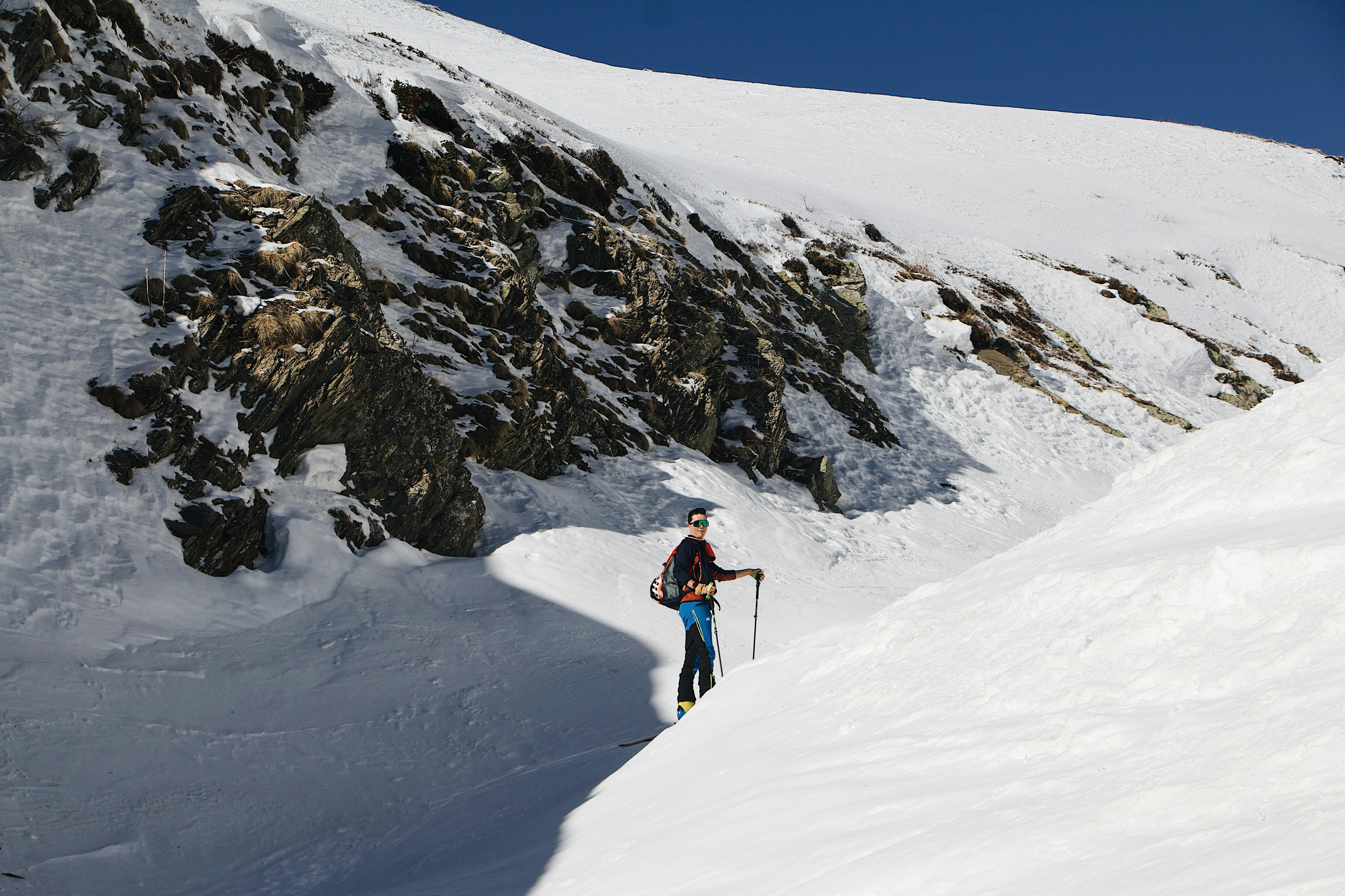 Skier traversing snowy mountain terrain with rocky slopes under a clear blue sky.