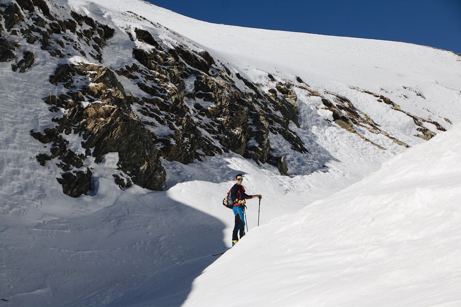 Skier traversing snowy mountain terrain with rocky slopes under a clear blue sky.