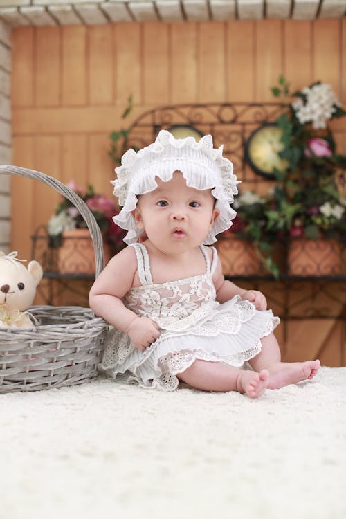 Gratis Bebé Con Diadema Blanca Y Vestido Floral De Encaje Blanco Sentado Junto A Una Canasta De Mimbre Gris Foto de stock