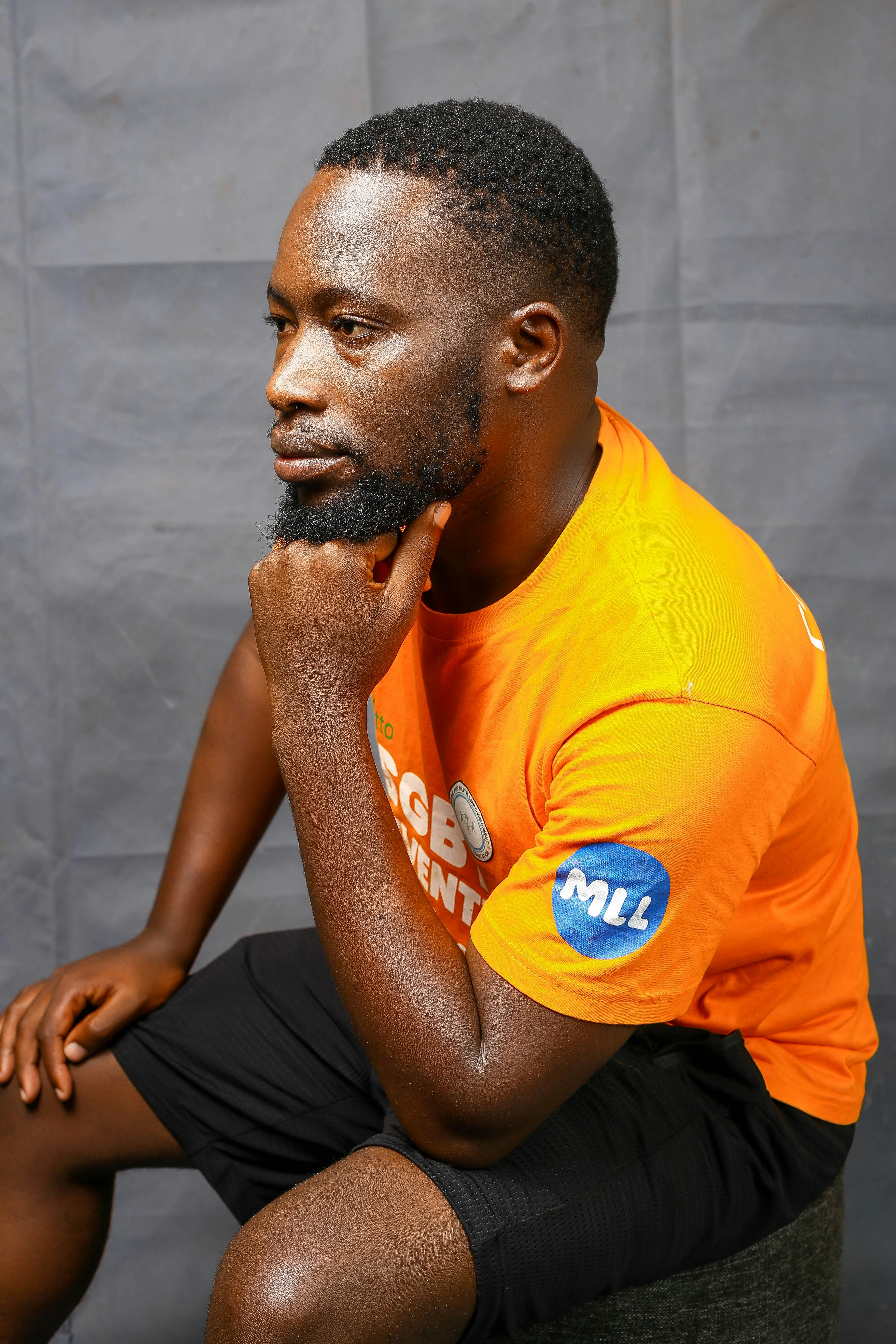 A thoughtful man sits indoors wearing a vibrant orange t-shirt, captured in a side profile portrait.
