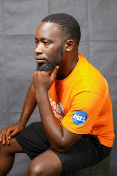 A thoughtful man sits indoors wearing a vibrant orange t-shirt, captured in a side profile portrait.