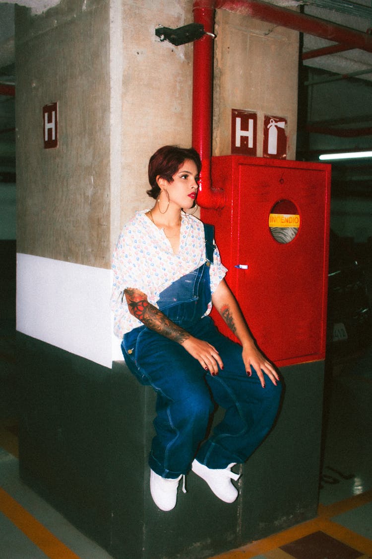 Young Woman With Tattoos Wearing Denim Overalls And Sitting On A Parking Lot 