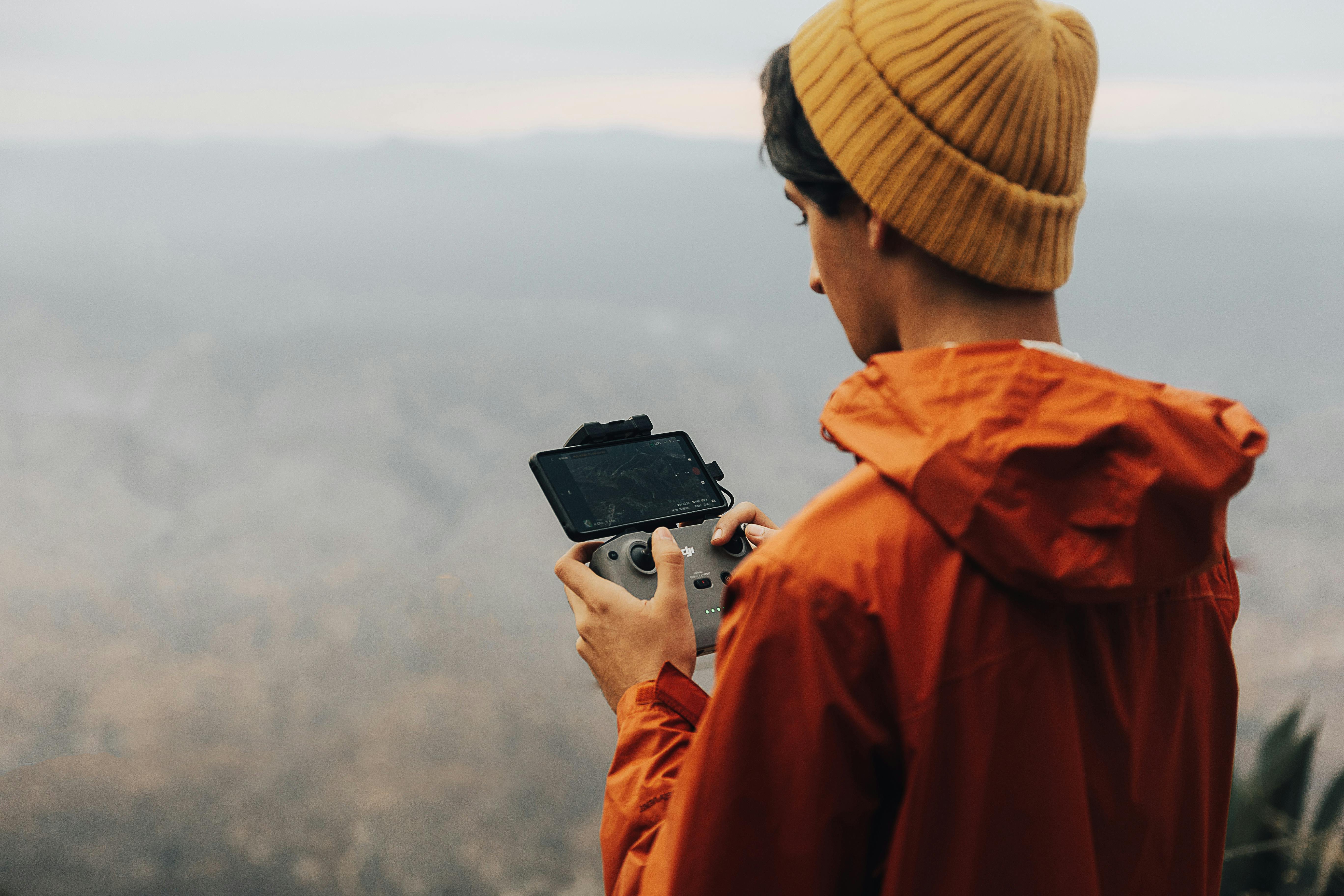 A young man in an orange jacket and beanie operates a drone controller in a foggy landscape.