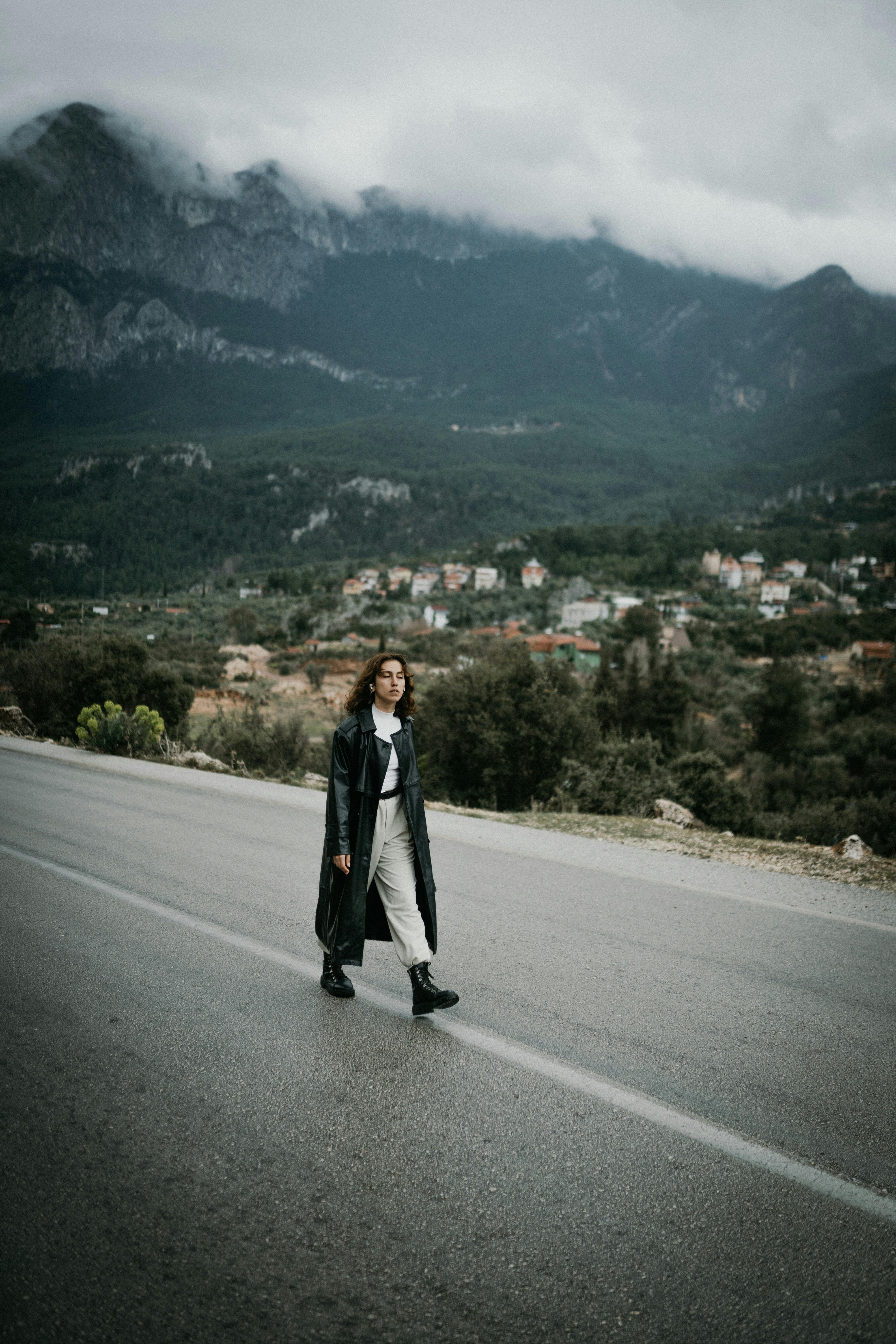A woman in a black coat walks on a mountain road with a village and mountains in the background.