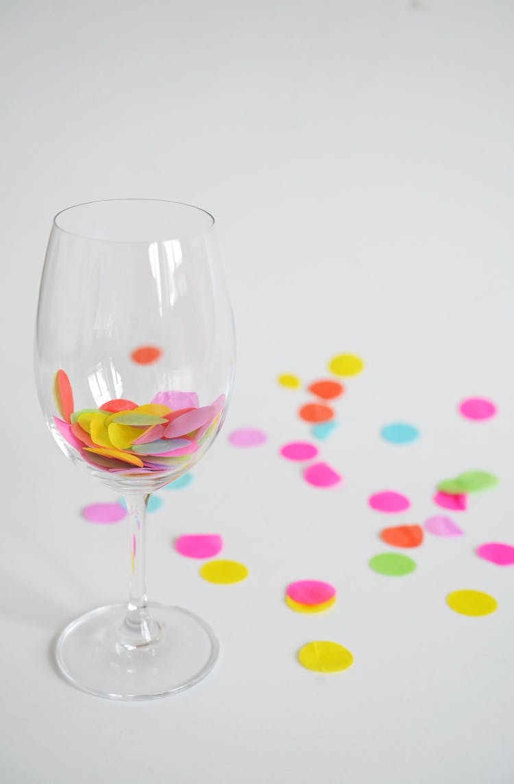 Colorful Confetti In A Wineglass And Scattered On White Surface
