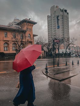 A person walking with a red umbrella on a rainy day in the city near modern architecture.