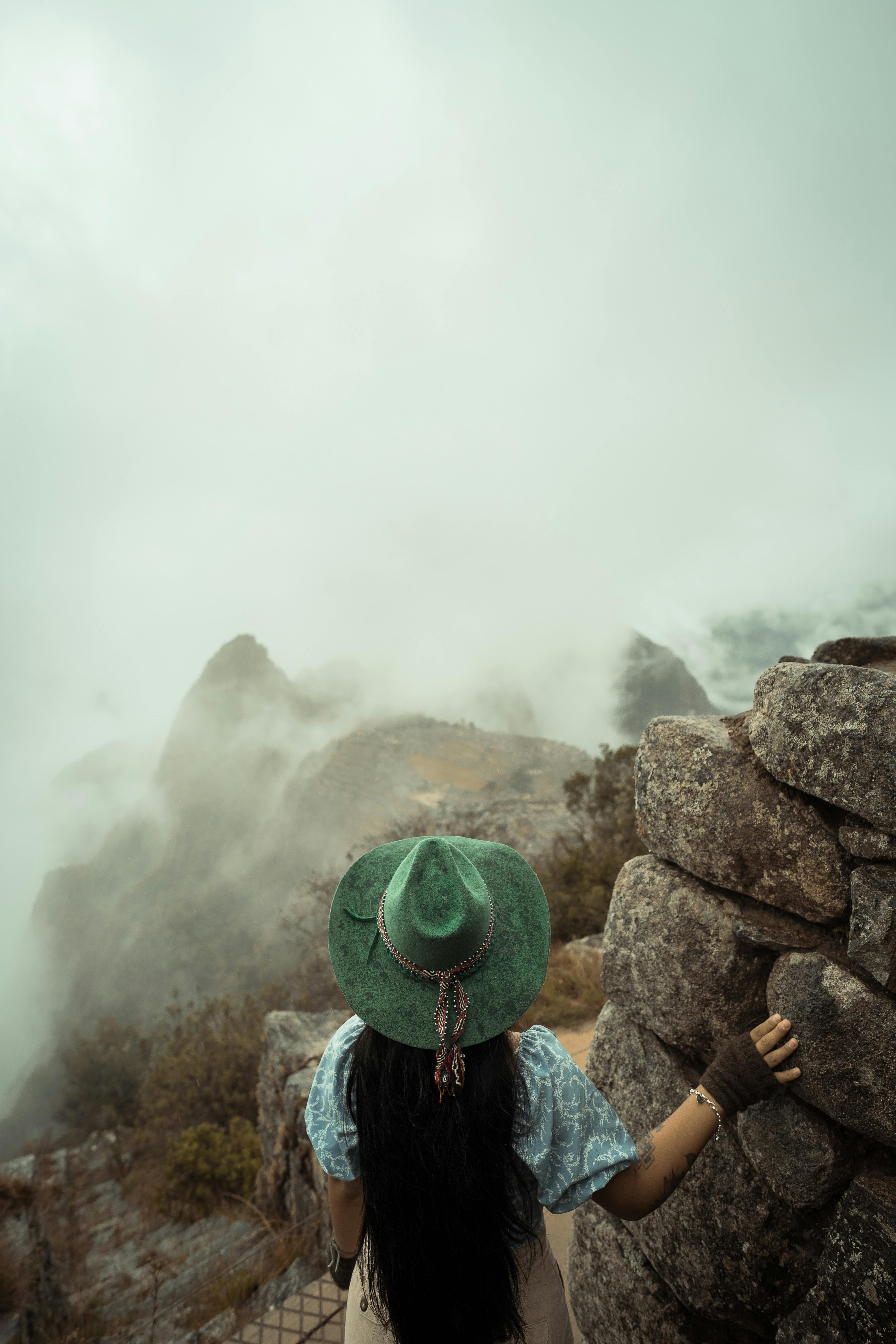 A woman in a green hat gazes over a misty mountain landscape in Cusco, Peru, surrounded by rocks and fog.