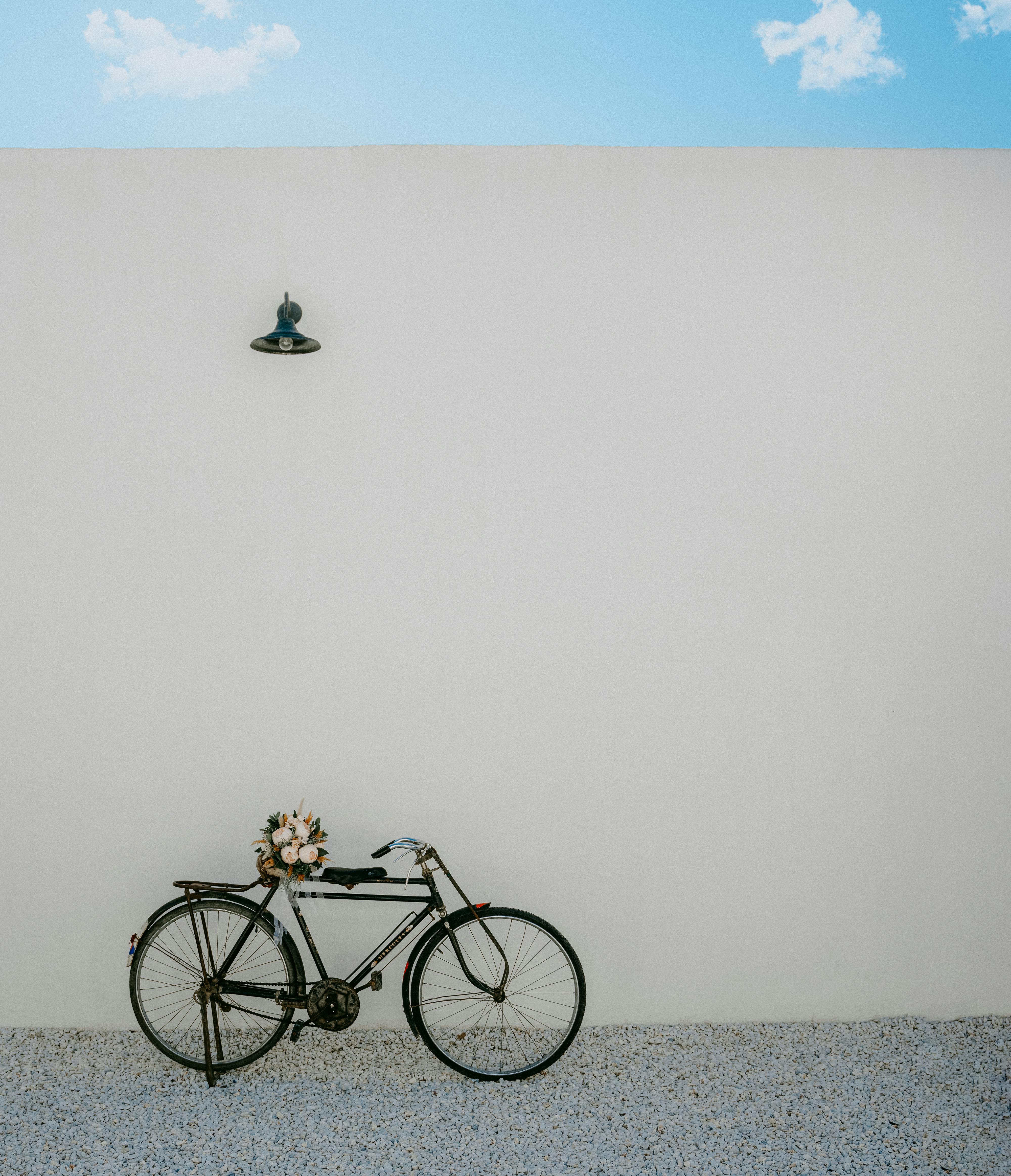 A rustic bicycle with a flower bouquet leans against a minimalist white wall under a clear sky.