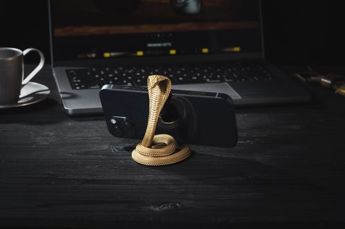 Free Golden cobra-shaped phone stand holding a smartphone, placed in front of a laptop on a dark surface. Stock Photo