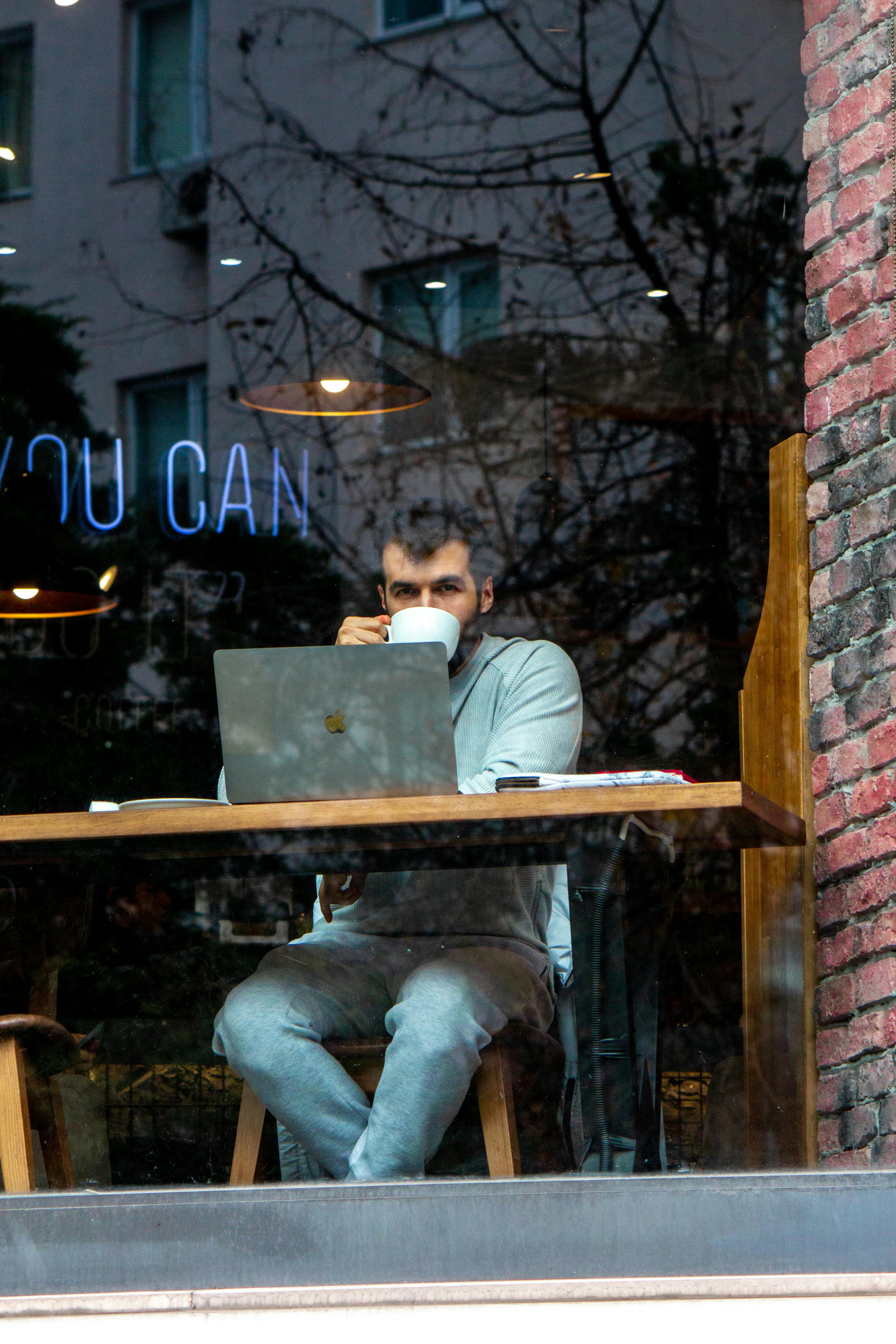 A man enjoys a coffee while working on his laptop in a cozy urban café, viewed from outside.