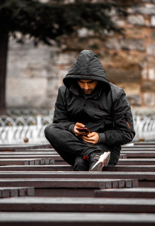 Free A man wearing a hooded jacket sits on a wooden bench outdoors using his smartphone. Stock Photo