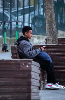Teenager with short hair sits on a park bench using a cellphone, surrounded by urban scenery.