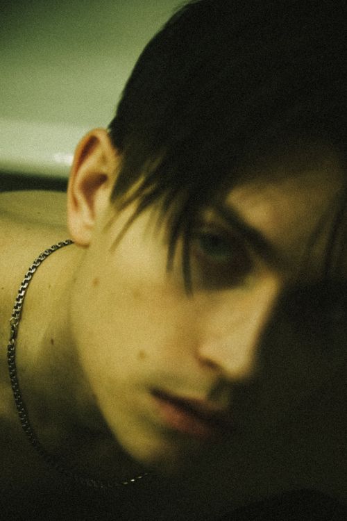 Free A close-up, moody portrait of a young man with dark hair wearing a chain necklace. Stock Photo