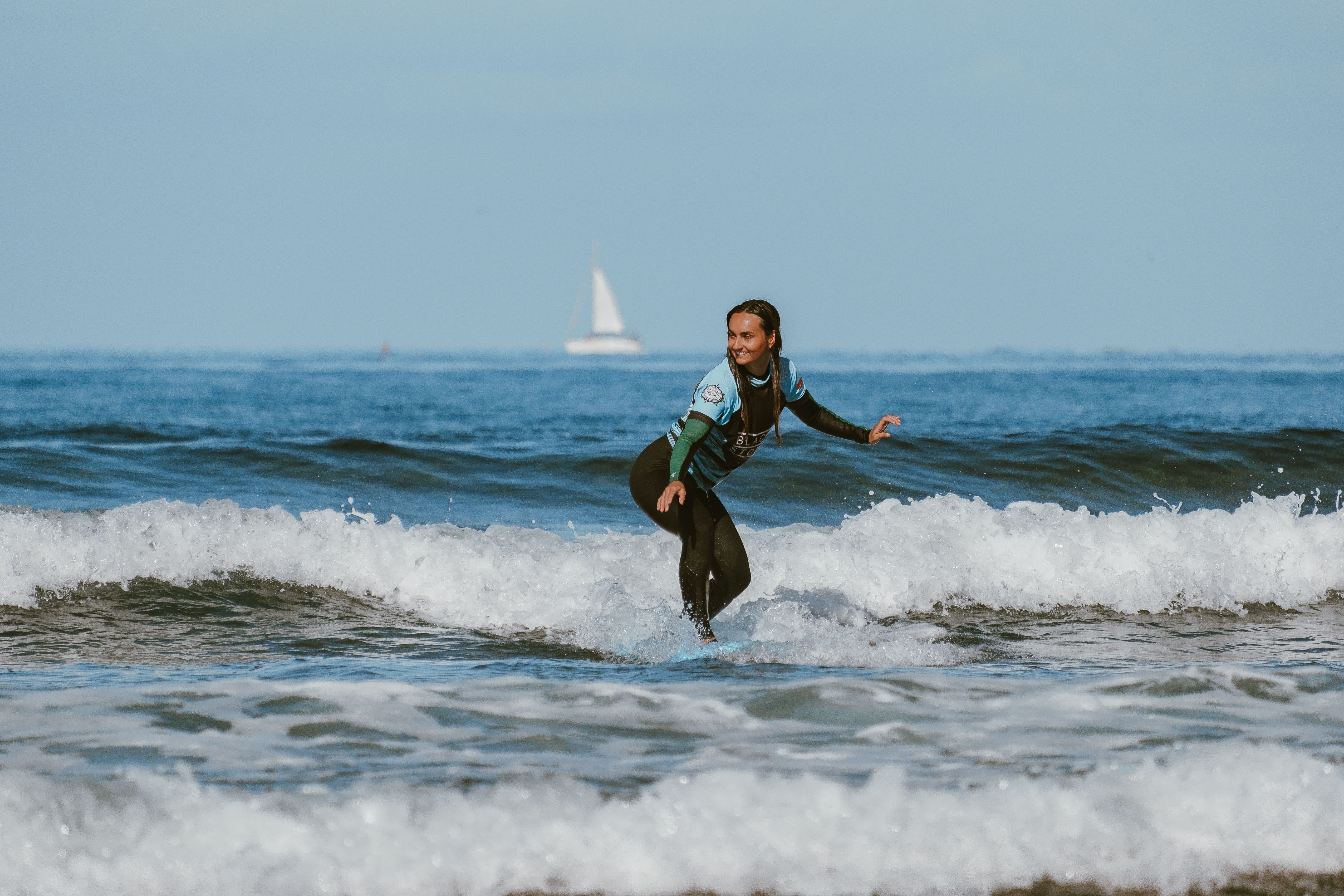 Energetic woman surfing on small ocean waves with a sailboat in the background.