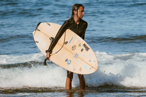 Free A woman with long hair holding a surfboard stands in ocean waves on a sunny day. Stock Photo