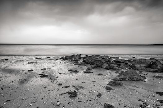 A serene black and white seascape with rocks and gentle waves on a sandy shore.