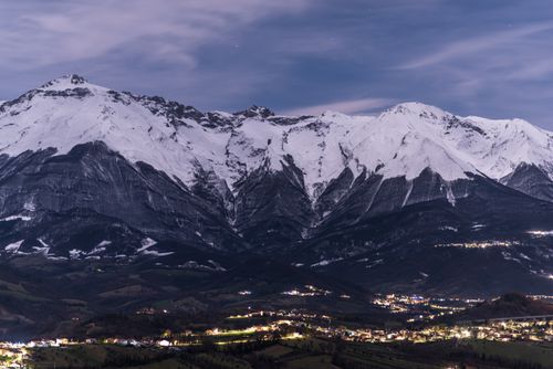 Free A serene snowy mountain range above a lit town during twilight, showcasing winter beauty. Stock Photo