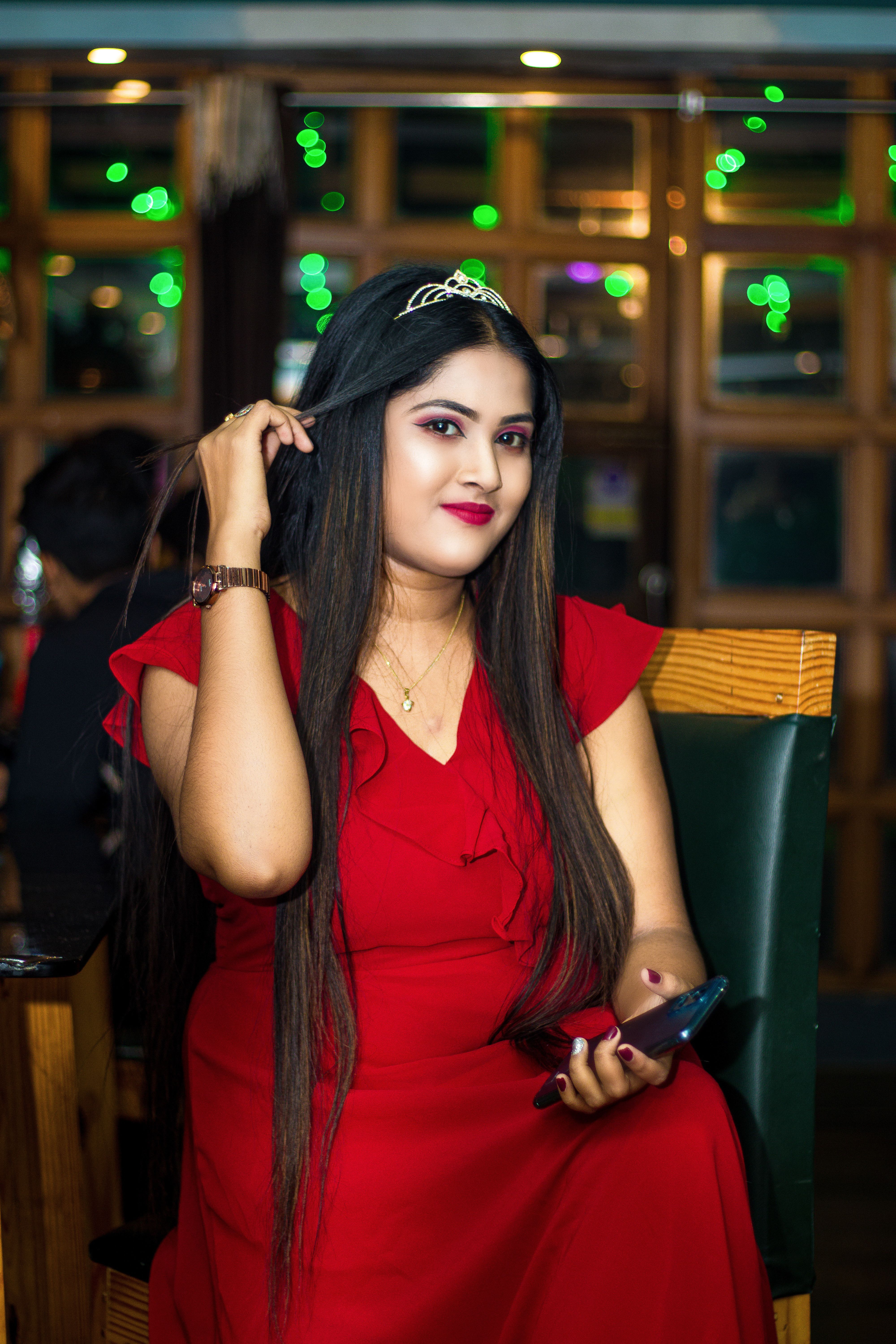 Portrait of a stylish woman in a red dress with a tiara, sitting indoors holding a smartphone.