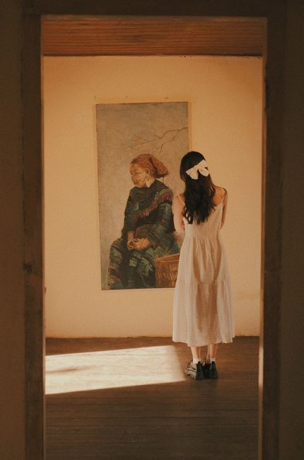 A woman stands in a gallery, admiring a painting on the wall. The scene is warmly lit and serene.