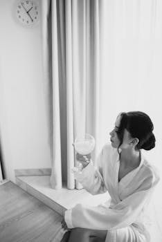Black and white photo of a woman in a bathrobe enjoying a drink by the window.
