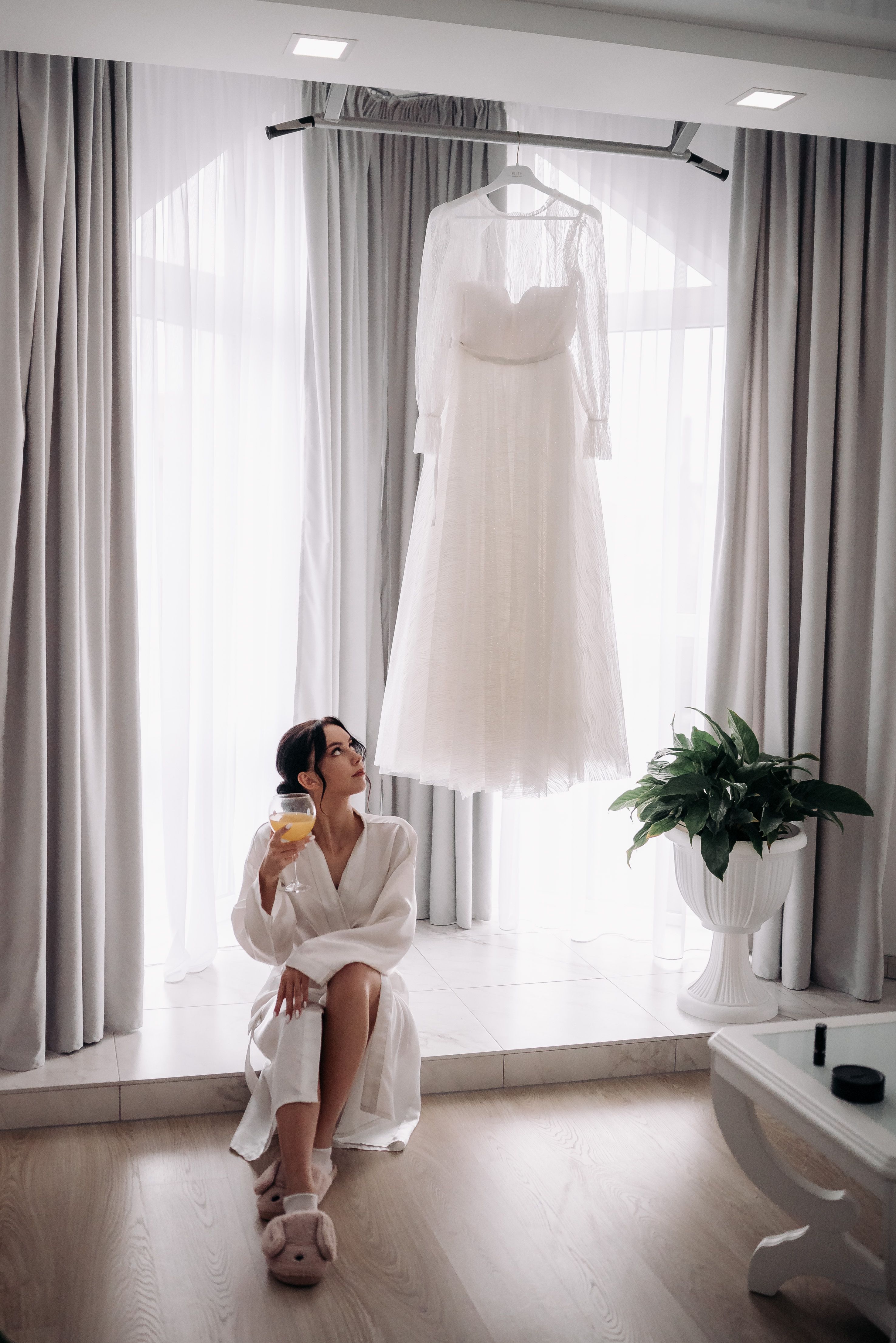Young woman in robe enjoys a drink while admiring her hanging wedding dress indoors.