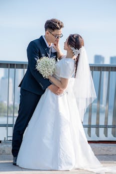 A couple in wedding attire embracing lovingly on a bridge with a cityscape backdrop.
