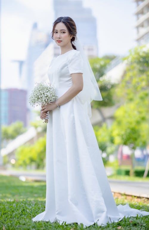Portrait of a Young Bride Standing on the Grass with a Bouquet of White Wildflowers in Hands
