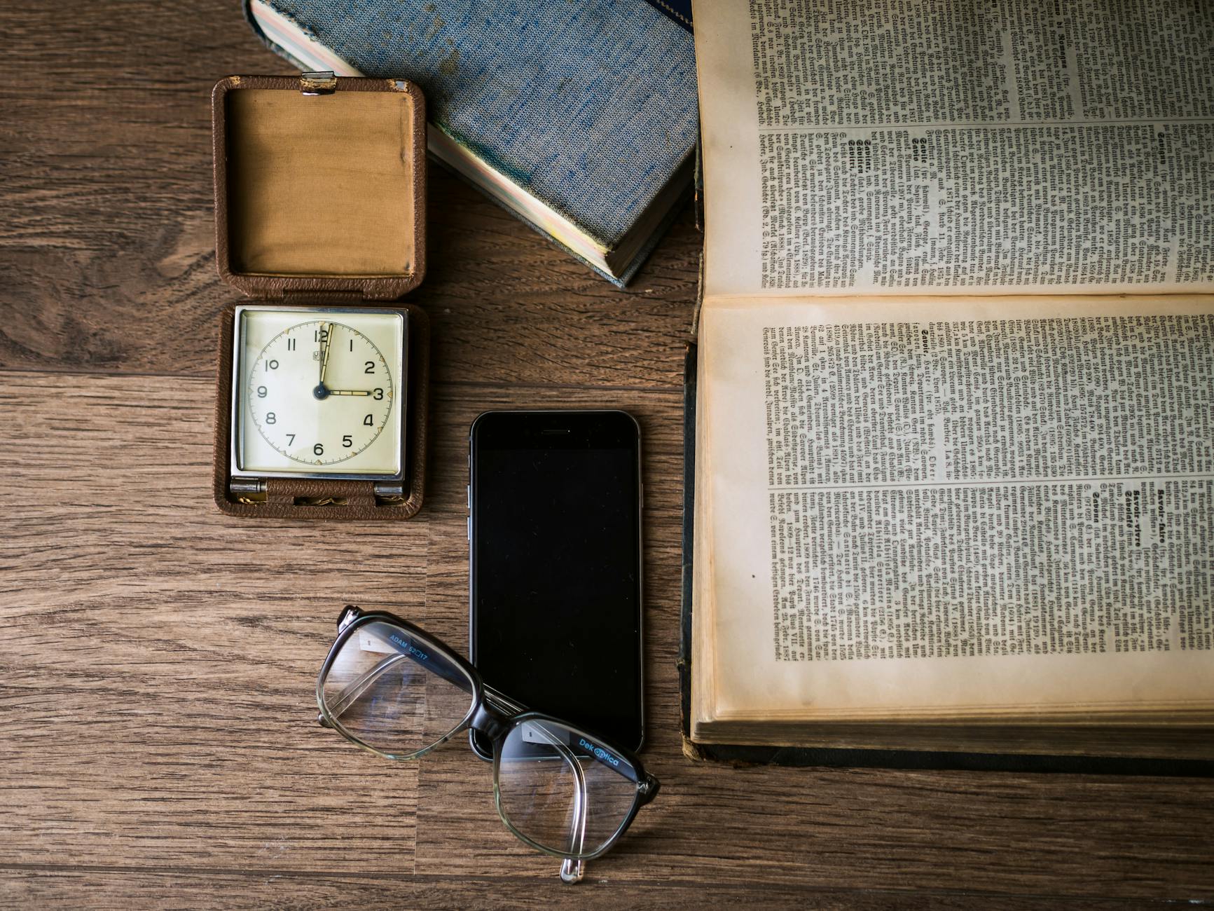 Flat lay of a vintage clock, open book, smartphone, and eyeglasses on a wooden table.