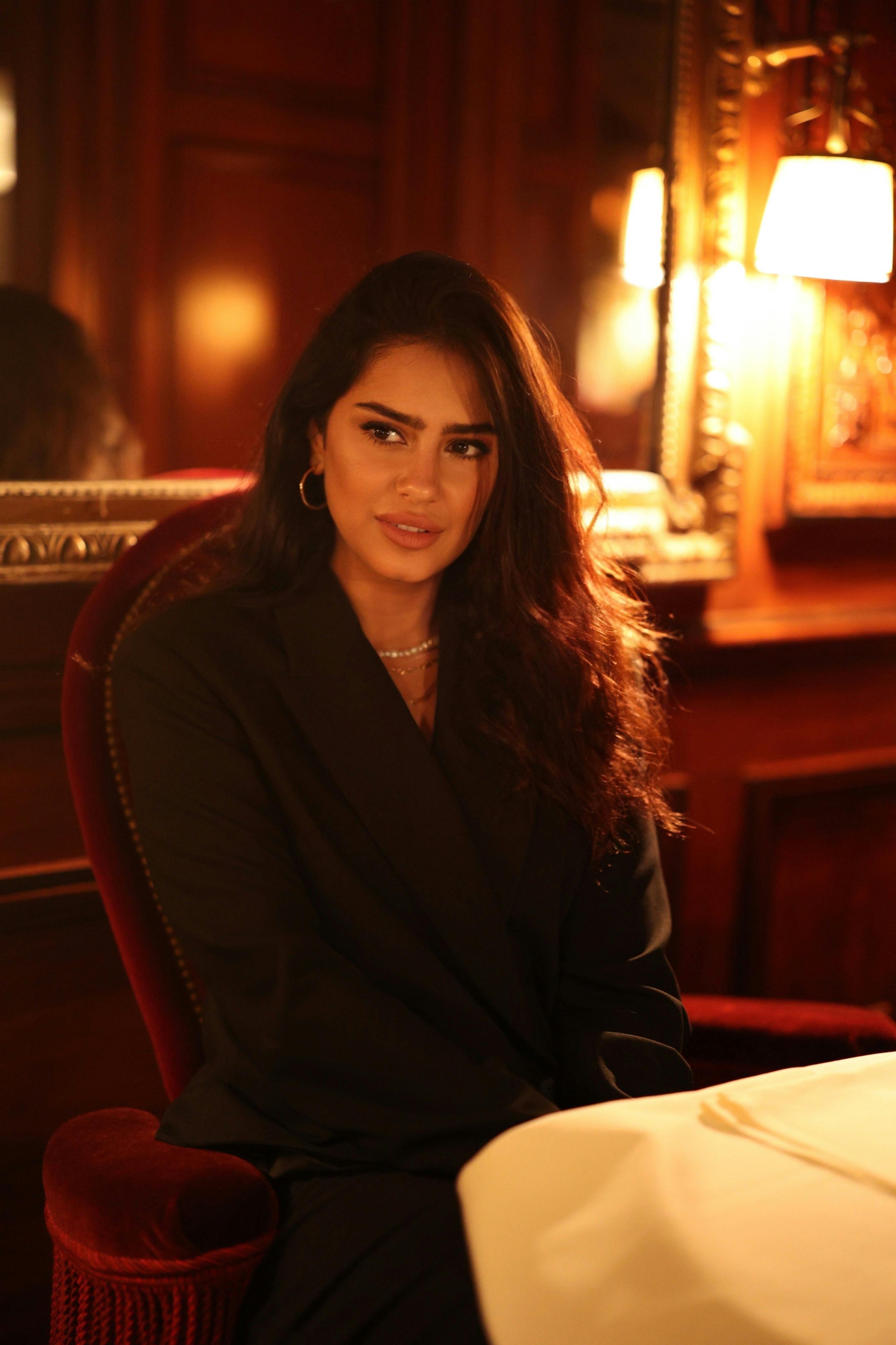 A young woman in a stylish outfit sits at a table in a warmly lit, elegant restaurant interior.